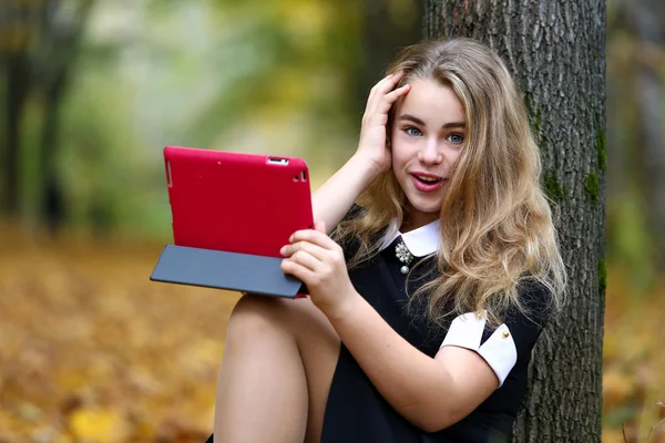 Colegiala sentada en el parque de otoño y sosteniendo una tabl digital — Foto de Stock