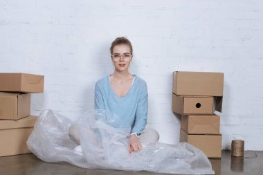 karton kutular arasında katta oturan ve kameraya bakarak gözlük genç girişimci