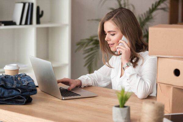 side view of young businesswoman talking on smartphone while working on laptop