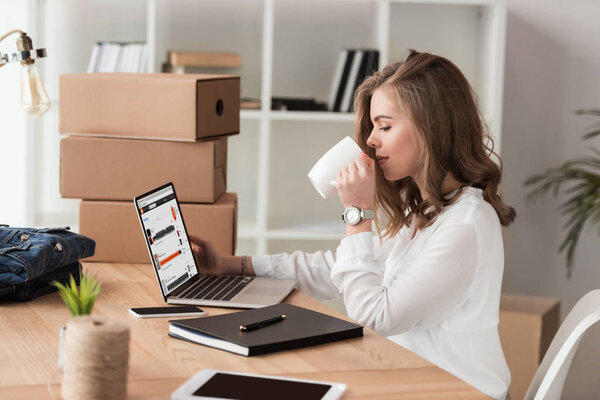 side view of businesswoman drinking coffee while working on laptop at table
