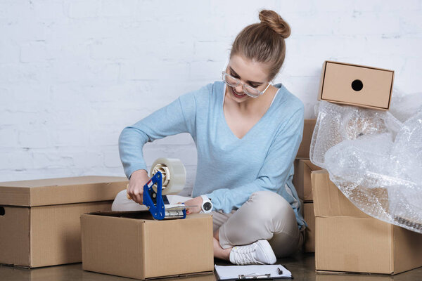 smiling entrepreneur packing parcel with adhesive tape at home office