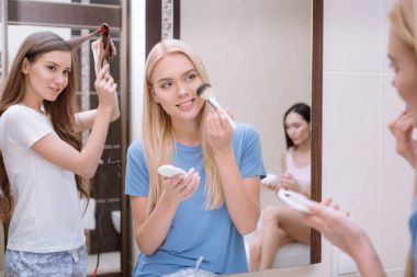 attractive girl applying foundation powder with makeup brush in bathroom with friends clipart