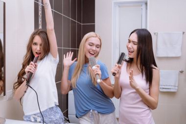 multiethnic friends singing in bathroom with round combs and hair iron  clipart