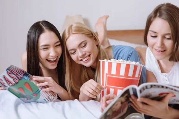 Smiling Multiethnic Girls Lying Bed Popcorn Reading Magazines — Free Stock Photo