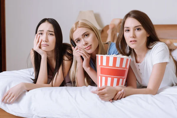 sad multiethnic girls lying on bed with popcorn and watching tv