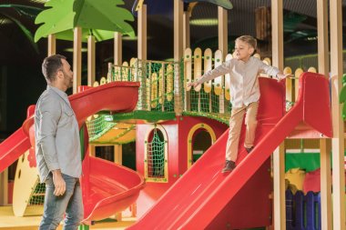 father looking at cute little son playing on slide in entertainment center clipart