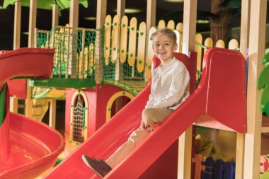 cute little boy smiling at camera while playing on slide at indoor play center clipart