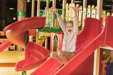 adorable happy little boy with raised hands playing on slide in game center clipart