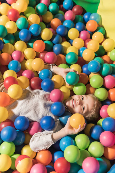 high angle view of cute little boy smiling at camera while lying in pool with colorful balls