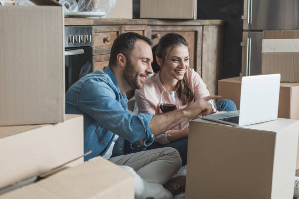 счастливая пара пьет вино и использует ноутбук в новом доме
