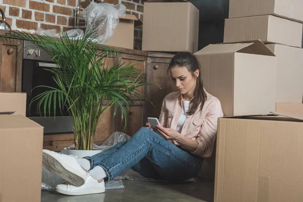 Νεαρή Γυναίκα Χρησιμοποιώντας Smartphone Καθμένος Μεταξύ Χαρτοκιβώτια Στο Νέο Διαμέρισμα — Φωτογραφία Αρχείου