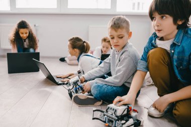 focused kids programming robots with laptops while sitting on floor, stem education concept clipart