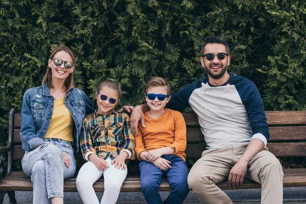 Familia sonriente en gafas de sol sentada en el banco - foto de stock
