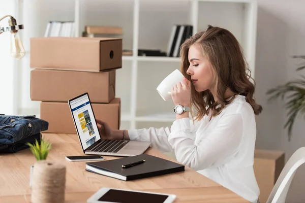 Vista lateral de la mujer de negocios beber café mientras trabaja en el ordenador portátil en la mesa - foto de stock