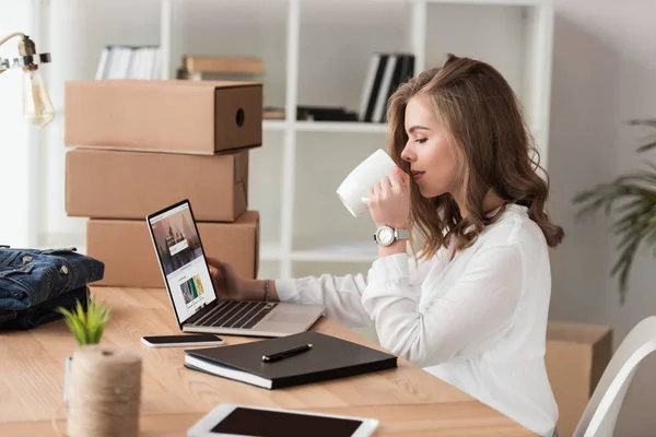 Vista lateral de empresária beber café enquanto trabalhava no laptop à mesa — Fotografia de Stock