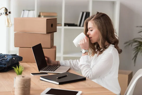 Vista lateral de empresária beber café enquanto trabalhava no laptop à mesa — Fotografia de Stock