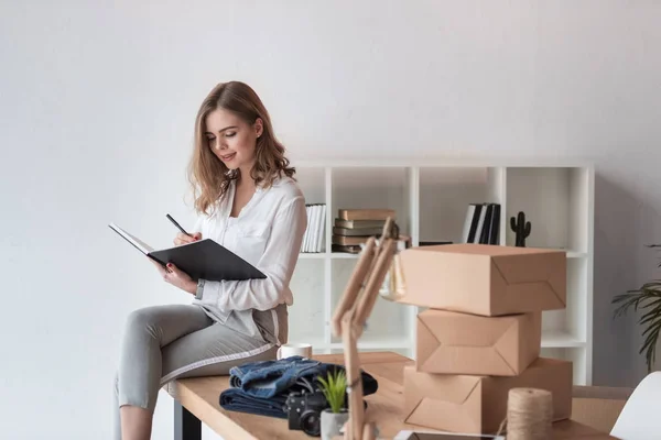 Jeune entrepreneur prenant des notes dans un carnet tout en étant assis sur la table au bureau à domicile — Photo de stock