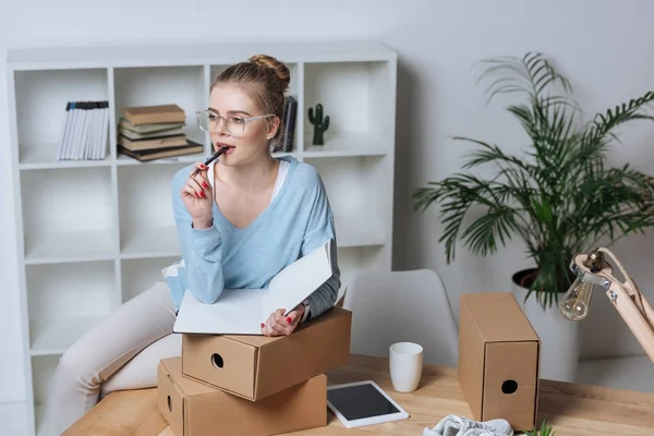 Porträt einer nachdenklichen Online-Shop-Inhaberin mit Stift und Notizbuch, die im Home Office wegschaut — Stockfoto