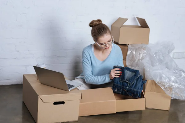 Молодий підприємець кладе джинсову куртку в картонну коробку під час упаковки продуктів в домашньому офісі — стокове фото