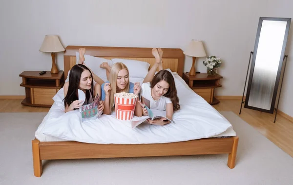 Chicas multiétnicas felices acostadas en la cama con palomitas de maíz y leyendo revistas - foto de stock