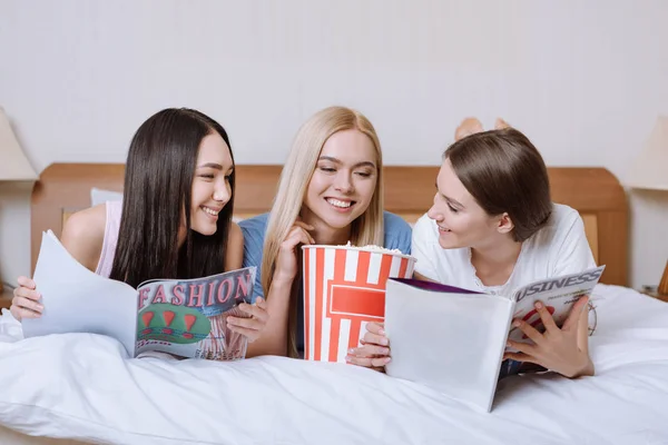 Multiethnic friends lying on bed with popcorn and reading magazines — Stock Photo