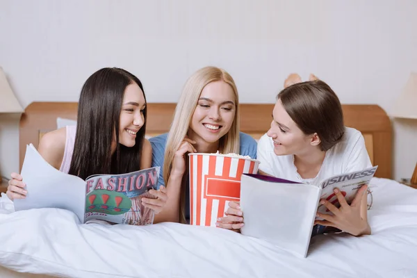 Amigos multiétnicos deitados na cama com pipocas e revistas de leitura — Fotografia de Stock