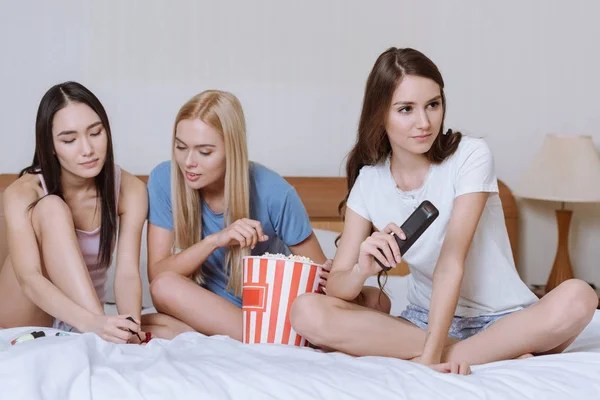 Multicultural girls sitting on bed with popcorn and watching tv — Stock Photo