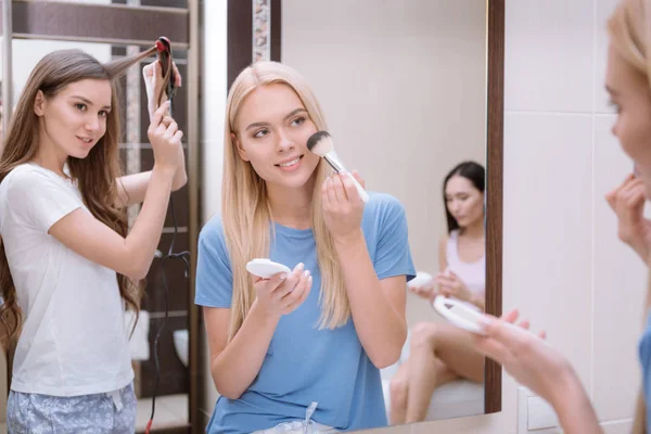 Menina atraente aplicando pó de fundação com escova de maquiagem no banheiro com amigos — Fotografia de Stock