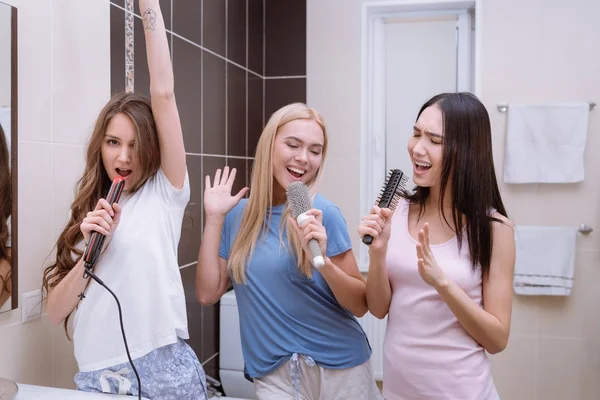 Многонациональные друзья поют в ванной комнате с круглыми расческами и утюгом для волос — стоковое фото