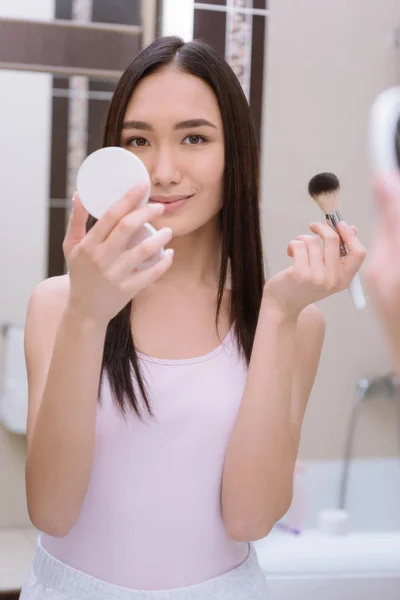 Asiático chica holding base polvo y maquillaje cepillo en cuarto de baño - foto de stock