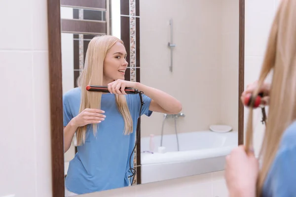 Chica sonriente alisando el cabello en el baño con plancha de pelo - foto de stock