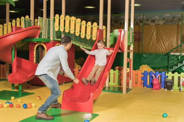Pai com adorável filho brincando no slide no centro de jogos indoor — Fotografia de Stock