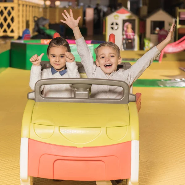 Adorabili fratelli felici seduti in macchina giocattolo e sorridenti alla fotocamera nel centro di intrattenimento — Foto stock