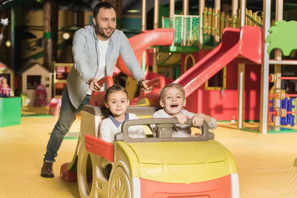 Padre sorridente che porta adorabili bambini felici sulla macchina giocattolo al centro di intrattenimento — Foto stock