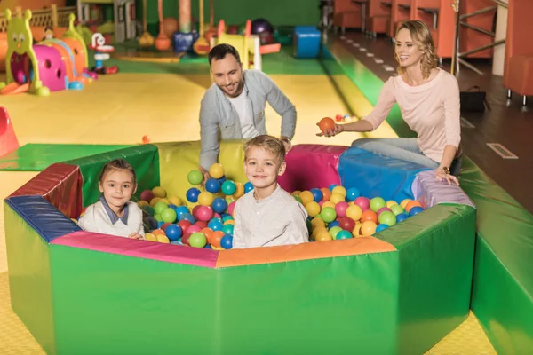 Щасливі батьки дивляться на милих маленьких дітей, посміхаючись на камеру, сидячи в басейні з різнокольоровими кульками — стокове фото