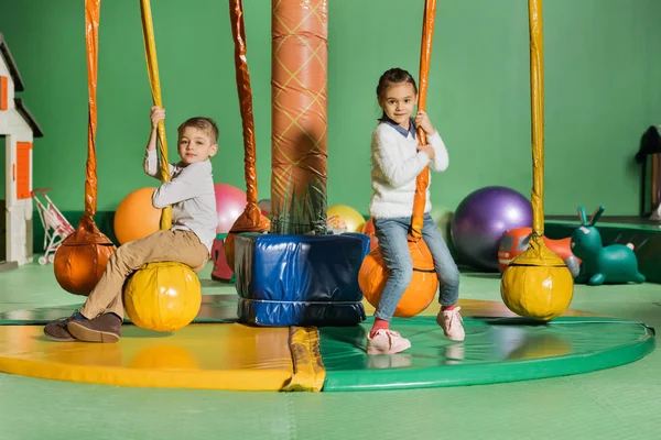 Süße glückliche kleine Kinder, die in die Kamera schauen, während sie im Unterhaltungszentrum schaukeln — Stockfoto