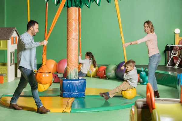 Pais sorridentes olhando para crianças felizes balançando em balanços no centro de jogos indoor — Fotografia de Stock