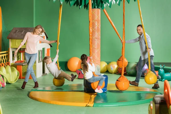 Pais sorridentes olhando para crianças felizes balançando em balanços no centro de entretenimento — Fotografia de Stock