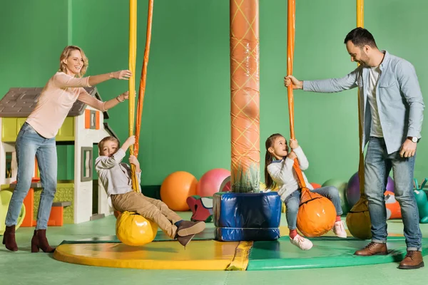 Glückliche Eltern, die Kinder auf Schaukeln im Vergnügungszentrum schaukeln sehen — Stockfoto