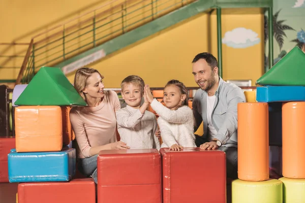 Padres felices mirando a los niños dando los cinco altos y sonriendo a la cámara en el centro de entretenimiento - foto de stock