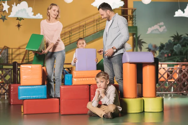 Bored little boy sitting on floor while happy family playing with blocks in entertainment center — Stock Photo