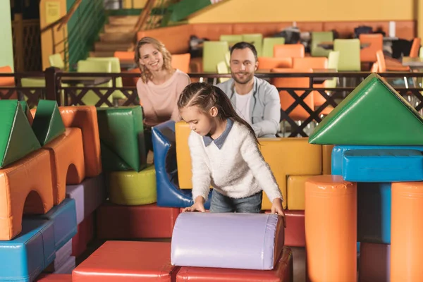 Eltern schauen sich niedliche kleine Tochter an, die im Unterhaltungszentrum ein Schloss mit bunten Bauklötzen baut — Stockfoto