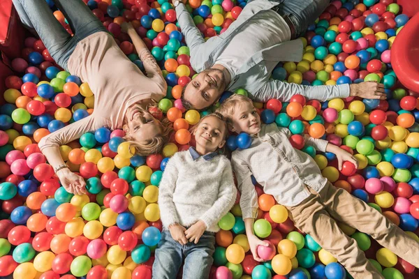 Вид зверху на щасливу сім'ю з двома дітьми, посміхаючись на камеру, лежачи в басейні з різнокольоровими кульками — стокове фото