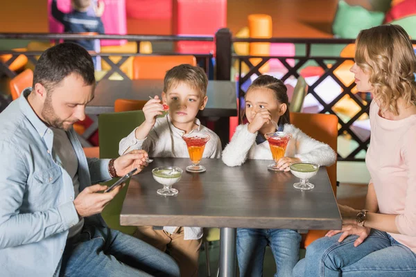 Familia con dos niños comiendo sabrosos postres y padre usando teléfono inteligente en el centro de entretenimiento - foto de stock