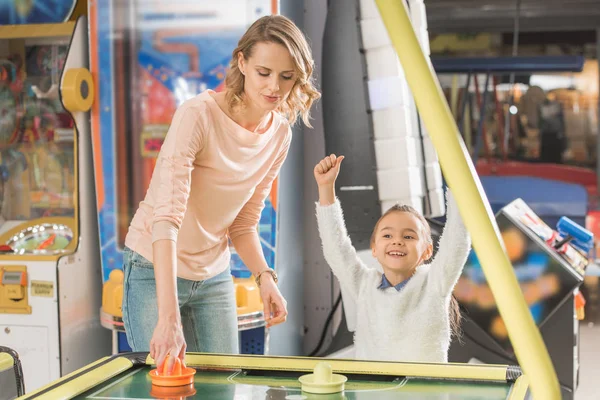 Sonriente madre y triunfante hijita jugando al hockey aéreo en el centro del juego - foto de stock