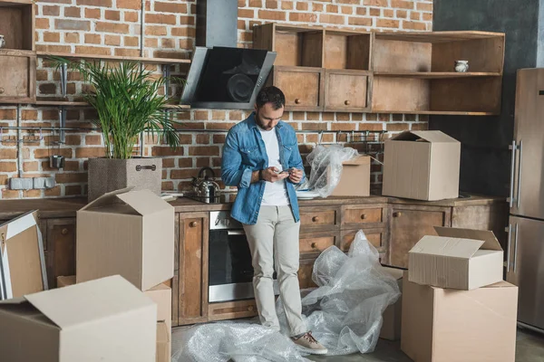 Hombre usando smartphone mientras está de pie entre cajas de cartón en nuevo apartamento - foto de stock