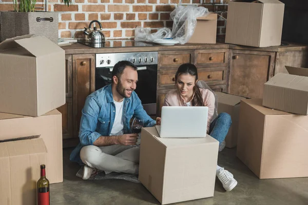 Lächelndes junges Paar trinkt Wein und benutzt Laptop in neuer Wohnung — Stockfoto