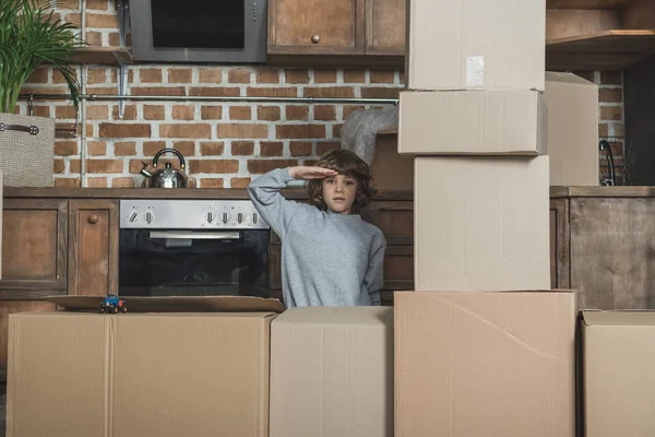 Niño pequeño saludando y mirando a la cámara mientras juega con cajas de cartón en nuevo apartamento - foto de stock