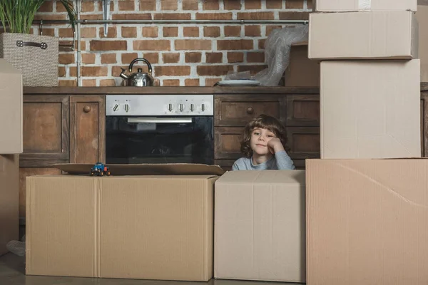 Cute little boy looking at camera while playing with cardboard boxes in new house — Stock Photo