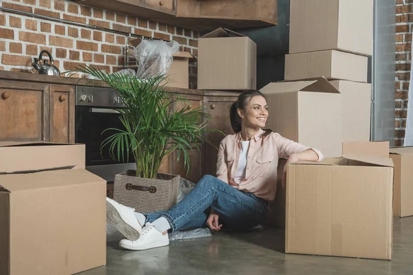 Feliz joven mujer sonriendo y mirando hacia otro lado mientras se sienta con cajas de cartón en nuevo apartamento - foto de stock